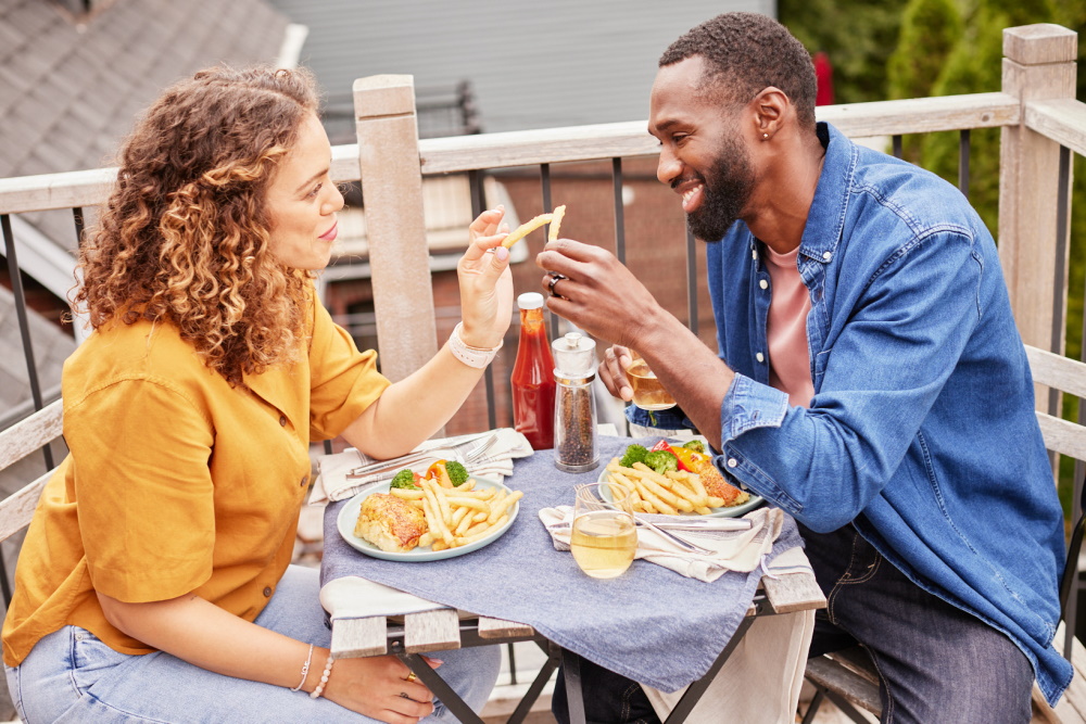 Couple assis à une table style bistro à deux places sur son patio levant leur verre à leur repas comprenant des frites. 